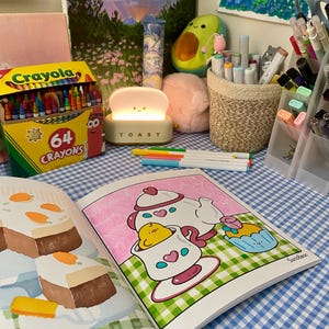 K&ouml;nnte beinhalten: Ein Stillleben mit einer Crayola-Buntstiftbox, einer originellen Lampe und einem offenen Malbuch. Die Szene umfasst Kunstzubeh&ouml;r, eine Avocado-Pl&uuml;schfigur und einen rosa Pompon auf einer blau-wei&szlig; karierten Oberfl&auml;che. Die Lampe sagt "TOAST".