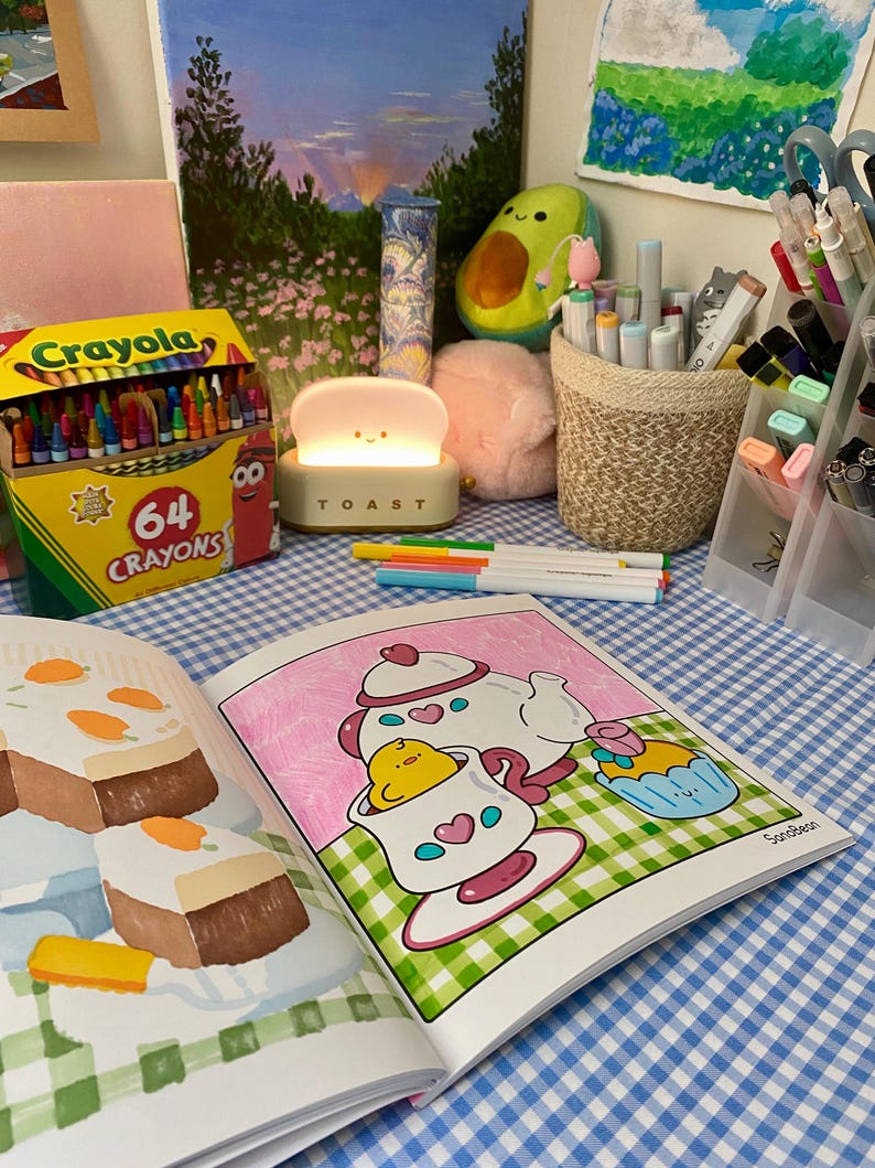 K&ouml;nnte beinhalten: Ein Stillleben mit einer Crayola-Buntstiftbox, einer originellen Lampe und einem offenen Malbuch. Die Szene umfasst Kunstzubeh&ouml;r, eine Avocado-Pl&uuml;schfigur und einen rosa Pompon auf einer blau-wei&szlig; karierten Oberfl&auml;che. Die Lampe sagt "TOAST".