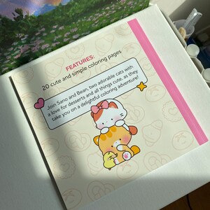 K&ouml;nnte beinhalten: Ein Malbuch mit einem rosa Rand und dem Text "FEATURES: 20 s&uuml;&szlig;e und einfache Malvorlagen". Das Buch zeigt Illustrationen von zwei Katzen und einem K&uuml;ken. Der Text lautet: "Begleite Sano und Bean, zwei bezaubernde Katzen mit einer Vorliebe f&uuml;r Desserts und alles, was s&uuml;&szlig; ist, auf ein entz&uuml;ckendes Malabenteuer!"