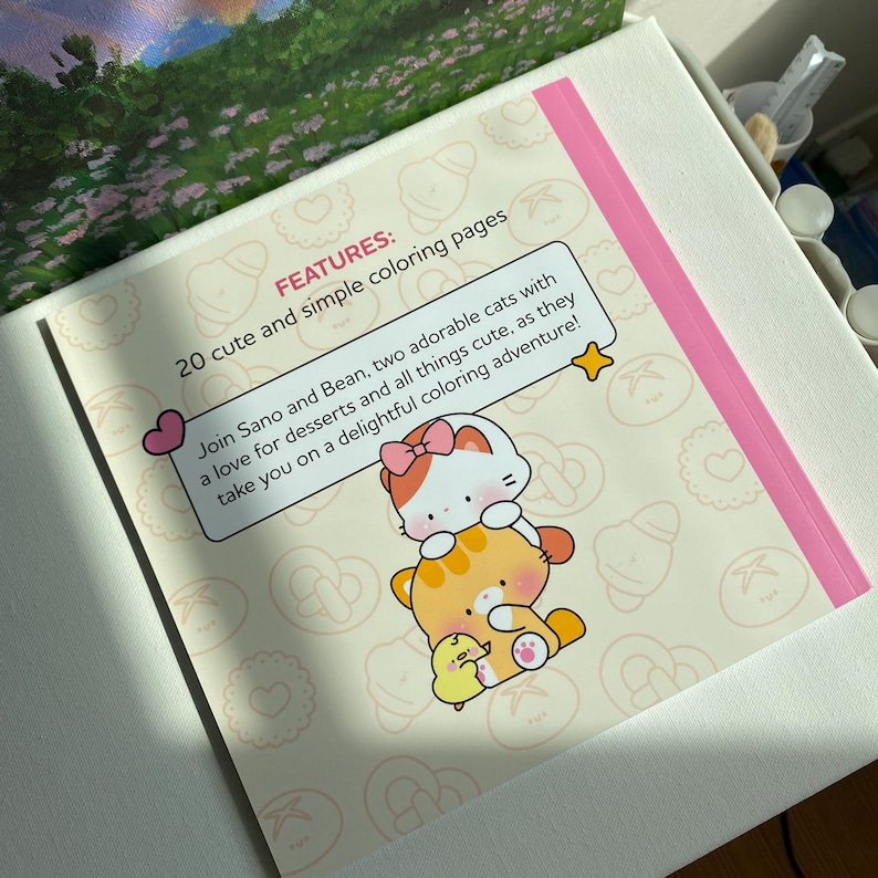 K&ouml;nnte beinhalten: Ein Malbuch mit einem rosa Rand und dem Text "FEATURES: 20 s&uuml;&szlig;e und einfache Malvorlagen". Das Buch zeigt Illustrationen von zwei Katzen und einem K&uuml;ken. Der Text lautet: "Begleite Sano und Bean, zwei bezaubernde Katzen mit einer Vorliebe f&uuml;r Desserts und alles, was s&uuml;&szlig; ist, auf ein entz&uuml;ckendes Malabenteuer!"