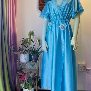 May include: A bright blue satin wrap-style robe featuring short flutter sleeves with delicate lace trim along the edges and neckline. The garment has an elasticized waist with a matching fabric tie closure and a tag attached to the front.