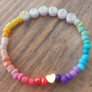 May include: A rainbow beaded bracelet with the name "MILLIE" spelled out in gold letters on white beads. A gold heart charm is in the centre of the bracelet.