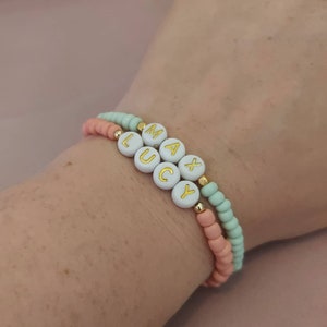 Puede incluir: Dos pulseras de cuentas con los nombres "MAX" y "LUCY" escritos en letras doradas sobre cuentas blancas. Las pulseras están superpuestas en la muñeca de una persona, con la pulsera superior con cuentas verde claro y la pulsera inferior con cuentas melocotón.
