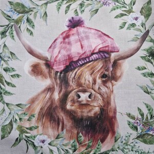 May include: A brown Highland cow wearing a pink and purple tartan hat with a white pom-pom. The cow is surrounded by a wreath of green leaves and white and purple flowers.