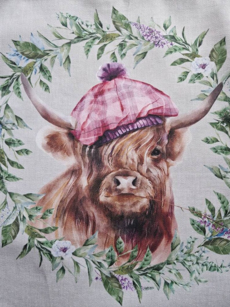 May include: A brown Highland cow wearing a pink and purple tartan hat with a white pom-pom. The cow is surrounded by a wreath of green leaves and white and purple flowers.