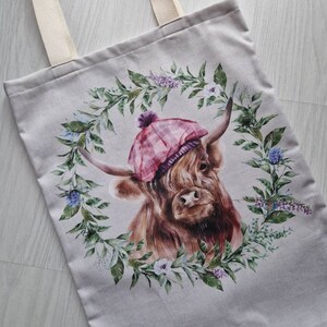 May include: A beige tote bag featuring a printed design of a Highland cow wearing a pink and white tartan hat. The cow is surrounded by a wreath of green leaves and flowers.
