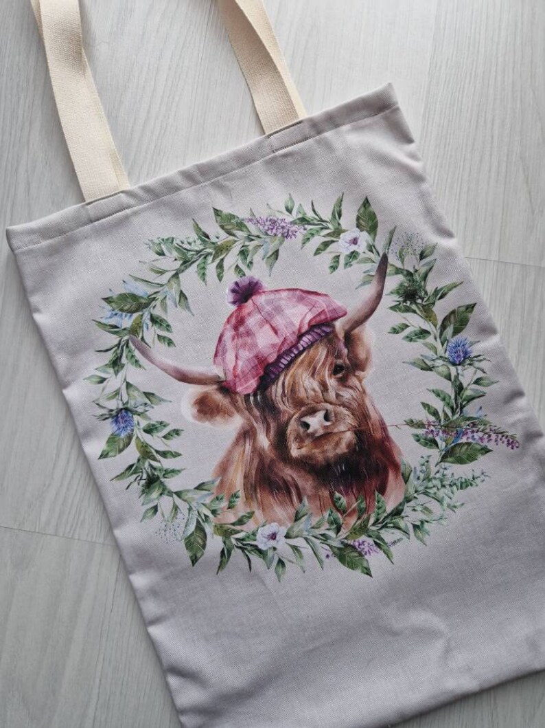 May include: A beige tote bag featuring a printed design of a Highland cow wearing a pink and white tartan hat. The cow is surrounded by a wreath of green leaves and flowers.
