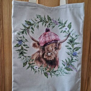 May include: A beige tote bag featuring a brown and white Highland cow wearing a pink and red tartan hat. The cow is surrounded by a wreath of green leaves and white flowers.