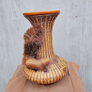 May include: A woven wicker vase with a brown and tan striped pattern. The vase has a squirrel figurine attached to the side. The squirrel is made of wood and has a brown and tan color.