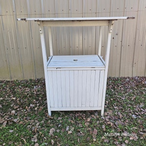 May include: White wooden outdoor bar cart with a slatted top and a storage compartment with a hinged lid. The cart is sitting on a bed of brown leaves.