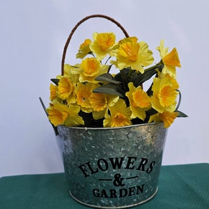 May include: A galvanized metal planter with a brown handle, filled with a bouquet of yellow daffodils. The planter has the text "FLOWERS & GARDEN" printed on the front.