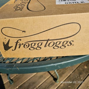 May include: A brown cardboard box with the brand name "frogg toggs" printed in black. The box is sitting on a green metal table. The date "November 03, 2025" is printed on the bottom right corner.