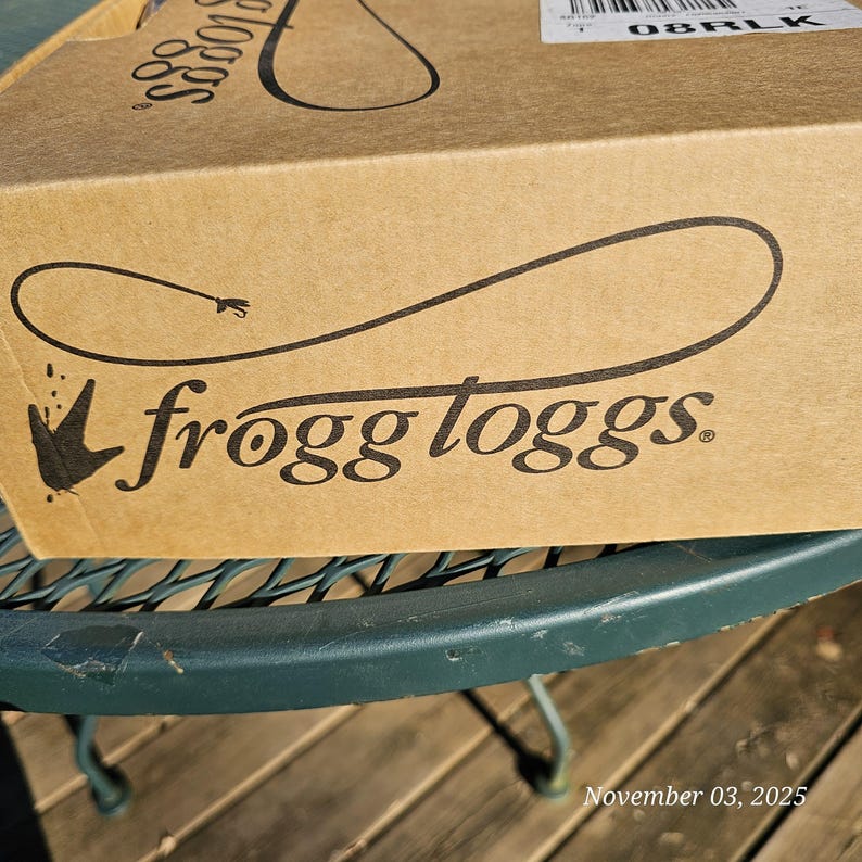 May include: A brown cardboard box with the brand name "frogg toggs" printed in black. The box is sitting on a green metal table. The date "November 03, 2025" is printed on the bottom right corner.