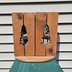 May include: A wooden decorative piece featuring two cut-out Christmas tree shapes. The natural wood has visible knots and a warm, reddish-brown hue. The piece is placed on a green surface, suggesting an outdoor or rustic theme.