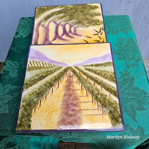May include: Two decorative ceramic plates with painted scenes. The top plate features a stylized depiction of olive trees with a yellow background. The bottom plate features a stylized depiction of a vineyard with a yellow and purple background. The text "Marilynn Bodway" is printed on the bottom plate.