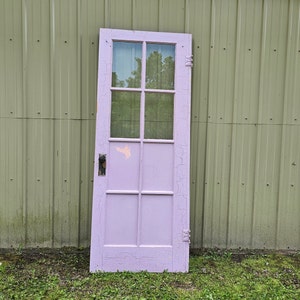 May include: A vintage, light purple wooden door with six glass panes. The door has a black door handle and is leaning against a green metal wall.