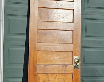 May include: A vintage wooden door with six rectangular panels and a brass doorknob. The door has a warm, natural wood tone with visible grain and is hinged on the left side. The door is leaning against a green garage door.