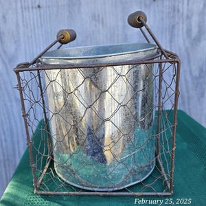 May include: A galvanized metal bucket with a wire mesh cage. The bucket has two handles with wooden grips. The wire mesh cage is rectangular and has a rustic, vintage appearance. The bucket is sitting on a green surface.