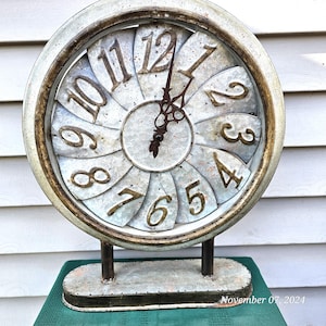 May include: A white metal clock with gold numbers and a distressed finish. The clock is shaped like a flower with petals that form the numbers. The clock is on a stand with a white metal base.