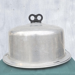 May include: A vintage aluminum cake carrier with a domed lid and a black handle. The carrier sits on a round base and is designed to protect and transport baked goods. The metal has a brushed finish, giving it a classic, retro look.