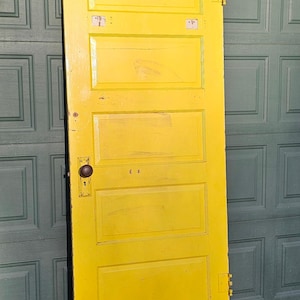 May include: A bright yellow, vintage wooden door with five rectangular panels. The door has a dark, round doorknob and metal hinges. The door is leaning against a green garage door.
