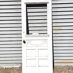 May include: A weathered, white wooden door with a decorative top panel and four lower panels. The door has a dark knob and is set against a gray, horizontal-paneled wall. The door is standing on dry grass.