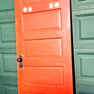 May include: A vibrant, orange-red wooden door with five rectangular panels. The door features a dark, round doorknob, hinges, and a latch. The door is leaning against a green garage door.