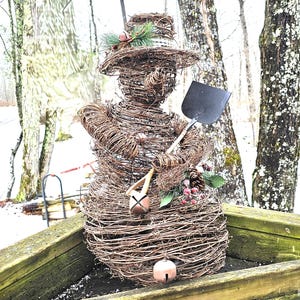 May include: A handcrafted snowman sculpture made of woven twigs, wearing a hat and holding a small shovel. The snowman is adorned with pinecones, berries, and bells. The sculpture is displayed outdoors, with a snowy backdrop and trees.