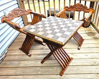 May include: A wooden chess table with two matching chairs. The table has a checkered surface and a decorative border. The chairs have ornate carved backs and armrests. The set is made of dark wood and is set on a wooden deck.