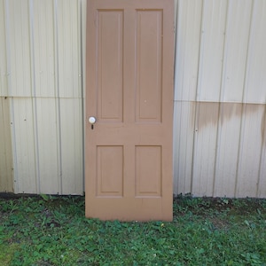 May include: A brown wooden door with four panels and a white door knob. The door is set against a white corrugated metal wall.