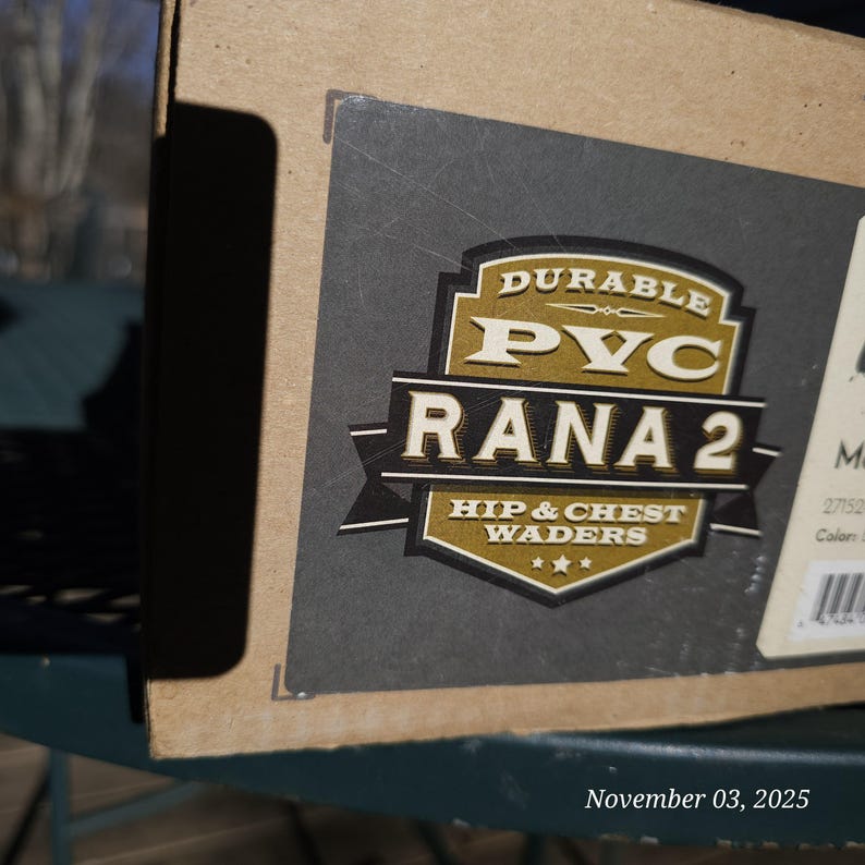 May include: A cardboard box with a label featuring the text "DURABLE PVC RANA 2 - HIP & CHEST WADERS." The label is primarily black and gold. The date "November 03, 2025" is printed at the bottom.