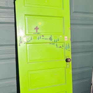 May include: A bright lime green door with a decorative landscape design. The door has a brass doorknob and hinges. The landscape design includes a cross, trees, and a sun. The door is approximately 80 inches tall.
