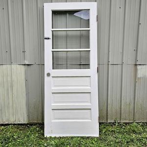 May include: A white, vintage door with a glass panel at the top, divided into four sections. The lower portion features three rectangular panels. The door has a handle and hinges, and is set against a corrugated metal background.