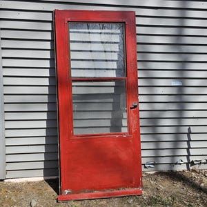 May include: A red wooden door with a single glass window pane. The door is on a gray siding house.