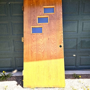 May include: Wooden door with three rectangular windows with blue inserts. The door is a light brown color with a yellow painted bottom half. The door is leaning against a dark green garage door.