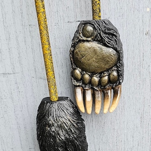 May include: Two decorative bear paw-shaped objects. The paws are detailed with black fur and gold claws. The paws are attached to yellow poles with black speckles. The background is a light gray wooden surface.