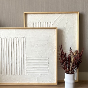 Puede incluir: Dos pinturas abstractas blancas texturizadas en marcos de madera, una encima de la otra, con un jarr&oacute;n blanco con flores secas delante de ellas.