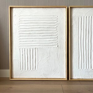 Puede incluir: Dos pinturas abstractas blancas texturizadas en marcos de madera. Las pinturas presentan l&iacute;neas en relieve y formas geom&eacute;tricas.