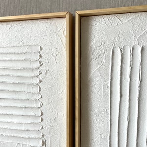 Puede incluir: Dos lienzos blancos texturizados con l&iacute;neas en relieve en un estilo abstracto moderno, enmarcados en madera natural.