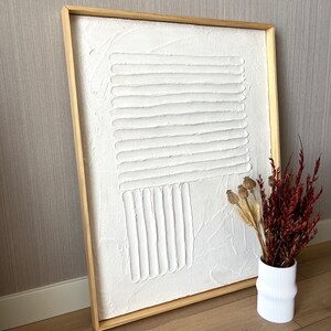 Puede incluir: Una pintura de lienzo blanco texturizado con un dise&ntilde;o geom&eacute;trico en relieve en un marco de madera natural.