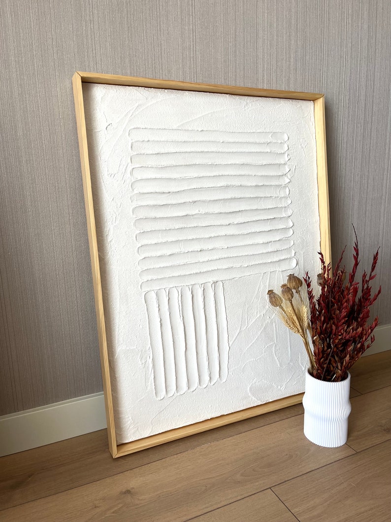 Puede incluir: Una pintura de lienzo blanco texturizado con un dise&ntilde;o geom&eacute;trico en relieve en un marco de madera natural.