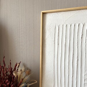 Puede incluir: Una pintura de lienzo blanco texturizado con un marco de madera. La pintura presenta una serie de l&iacute;neas verticales creadas con pintura blanca espesa.