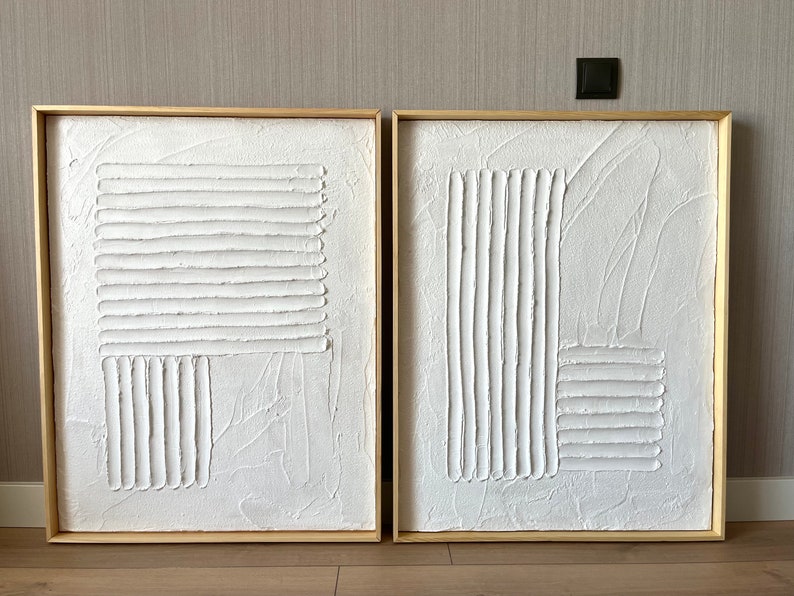 Puede incluir: Dos lienzos blancos texturizados con l&iacute;neas en relieve en un patr&oacute;n geom&eacute;trico, enmarcados en madera natural.