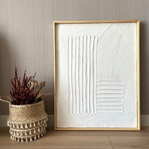 Puede incluir: Una pintura abstracta texturizada blanca en un marco de madera claro. La pintura presenta l&iacute;neas y formas en relieve, creando un dise&ntilde;o moderno y minimalista.