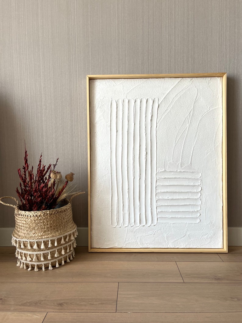 Puede incluir: Una pintura abstracta texturizada blanca en un marco de madera claro. La pintura presenta l&iacute;neas y formas en relieve, creando un dise&ntilde;o moderno y minimalista.