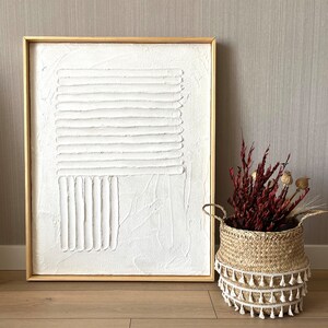 Puede incluir: Una pintura abstracta blanca texturizada en un marco de madera, con una cesta de mimbre natural con borlas blancas y flores secas rojas.