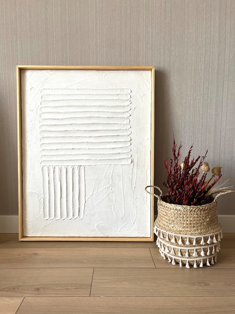 Puede incluir: Una pintura abstracta blanca texturizada en un marco de madera, con una cesta de mimbre natural con borlas blancas y flores secas rojas.