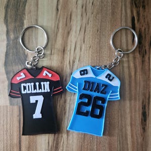 Op de afbeelding: Twee sleutelhangers in de vorm van een voetbalshirt. Eén is zwart met rode accenten, de naam "COLLIN" en het nummer "7". De andere is lichtblauw met witte accenten, de naam "DIAZ" en het nummer "26". Beide hebben zilveren sleutelringen.