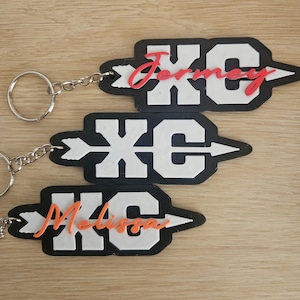 May include: Three black keychains with white "XC" lettering and arrow designs. The names "Jerney" and "Melissa" are in red and orange, respectively. Each keychain has a silver keyring and chain.