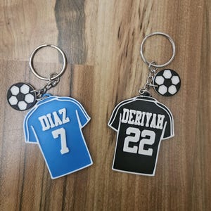 May include: Two soccer jersey keychains with a soccer ball charm. The blue jersey has the name "DIAZ" and the number "7" on the back. The black jersey has the name "DERIYAH" and the number "22" on the back.
